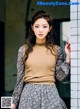 A woman leaning against a wall wearing a brown sweater and a floral skirt.