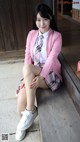 A woman in a pink blazer and plaid skirt sitting on a bench.