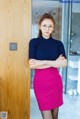 A woman in a blue sweater and pink skirt leaning against a wooden door.